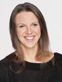 Porträt einer lächelnden Frau mit schulterlangem, glattem Haar. Sie trägt ein schwarzes Oberteil und steht vor einem hellen, neutralen Hintergrund. Ihr Lächeln wirkt herzlich und offen.