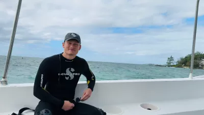 Ein Mann in Neoprenanzug sitzt auf einem Boot mit Blick auf das Meer, im Hintergrund Küste und teils bewölkter Himmel.