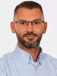 Ein Mann mit Brille und Bart trägt ein hellblaues Hemd und blickt freundlich in die Kamera, vor neutralem Hintergrund.