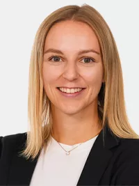 Eine junge Frau mit glattem, blondem Haar lächelt in die Kamera. Sie trägt ein dunkles Jackett über einem weißen Oberteil vor neutralem Hintergrund.