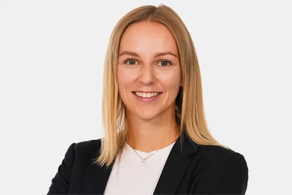 Eine junge Frau mit glattem, blondem Haar lächelt in die Kamera. Sie trägt ein dunkles Jackett über einem weißen Oberteil vor neutralem Hintergrund.