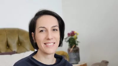 Eine lächelnde Person mit kurzen dunklen Haaren sitzt auf einem Sessel und hält eine Tasse in der Hand. Im Hintergrund stehen ein Strauß Blumen und ein Korb mit bunten Buchstaben.
