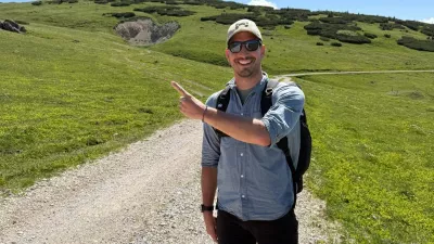 Mann mit Sonnenbrille, Rucksack und Kappe steht auf einem Schotterweg in den Bergen, lächelt und zeigt mit dem Finger auf den Hang im Hintergrund.