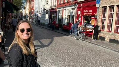 Eine lächelnde Frau steht auf einer sonnigen Kopfsteinpflasterstraße mit bunten Altstadthäusern im Hintergrund.