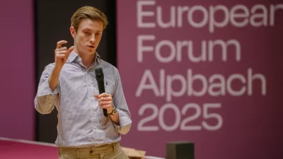 Ein junger Mann steht auf einer Bühne, hält ein Mikrofon in der Hand und gestikuliert beim Sprechen. Im Hintergrund ist eine große Leinwand mit der Aufschrift „European Forum Alpbach 2025“ zu sehen.