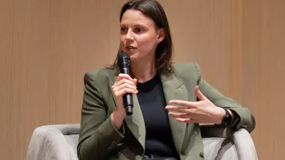 Nahaufnahme einer Frau, die auf einem Sessel sitzt und während einer Podiumsdiskussion in ein Mikrofon spricht. Sie trägt ein olivgrünes Sakko über einem dunklen Oberteil und gestikuliert mit der freien Hand vor einer hellen Holzpaneel-Wand.