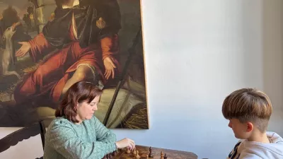 Ein Junge und eine Frau sitzen sich an einem kleinen Holztisch gegenüber und spielen konzentriert Schach. Die Frau trägt einen grünen Pullover und greift gerade nach einer Figur, während der Junge in einem hellen Hoodie aufmerksam zuschaut. Im Hintergrund hängt ein großes Gemälde mit einer religiösen Szene, das die ruhige, fast museale Atmosphäre des Raumes unterstreicht.