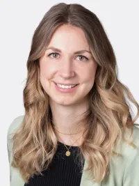 Porträt einer lächelnden Frau mit langen, welligen blond-braunen Haaren, hellem Blazer und schwarzem Oberteil vor neutralem Hintergrund.
