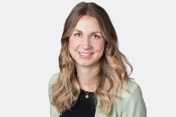 Porträt einer lächelnden Frau mit langen, welligen blond-braunen Haaren, hellem Blazer und schwarzem Oberteil vor neutralem Hintergrund.