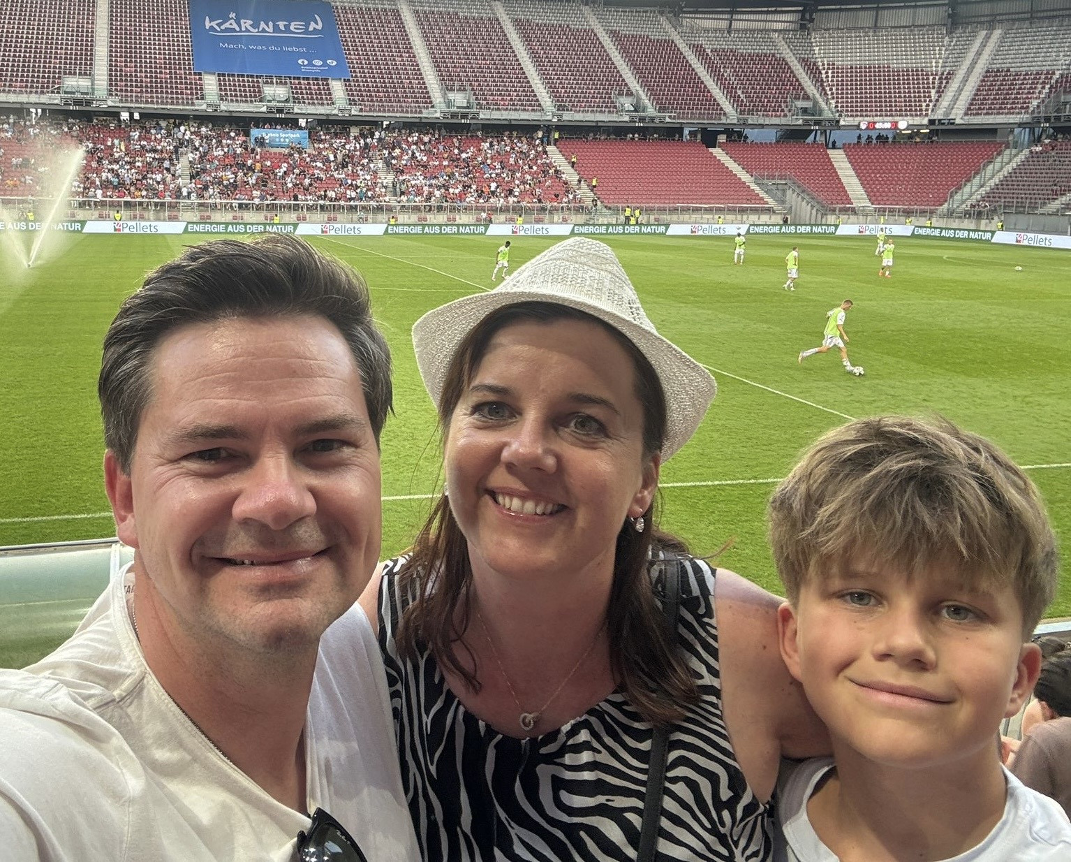 Drei Personen – ein Mann, eine Frau mit hellem Hut und ein Junge – machen ein Selfie in einem Fußballstadion und lächeln in die Kamera. Im Hintergrund ist das Spielfeld zu sehen, auf dem Spieler in Trainingskleidung stehen, sowie Tribünen mit vielen Zuschauern.