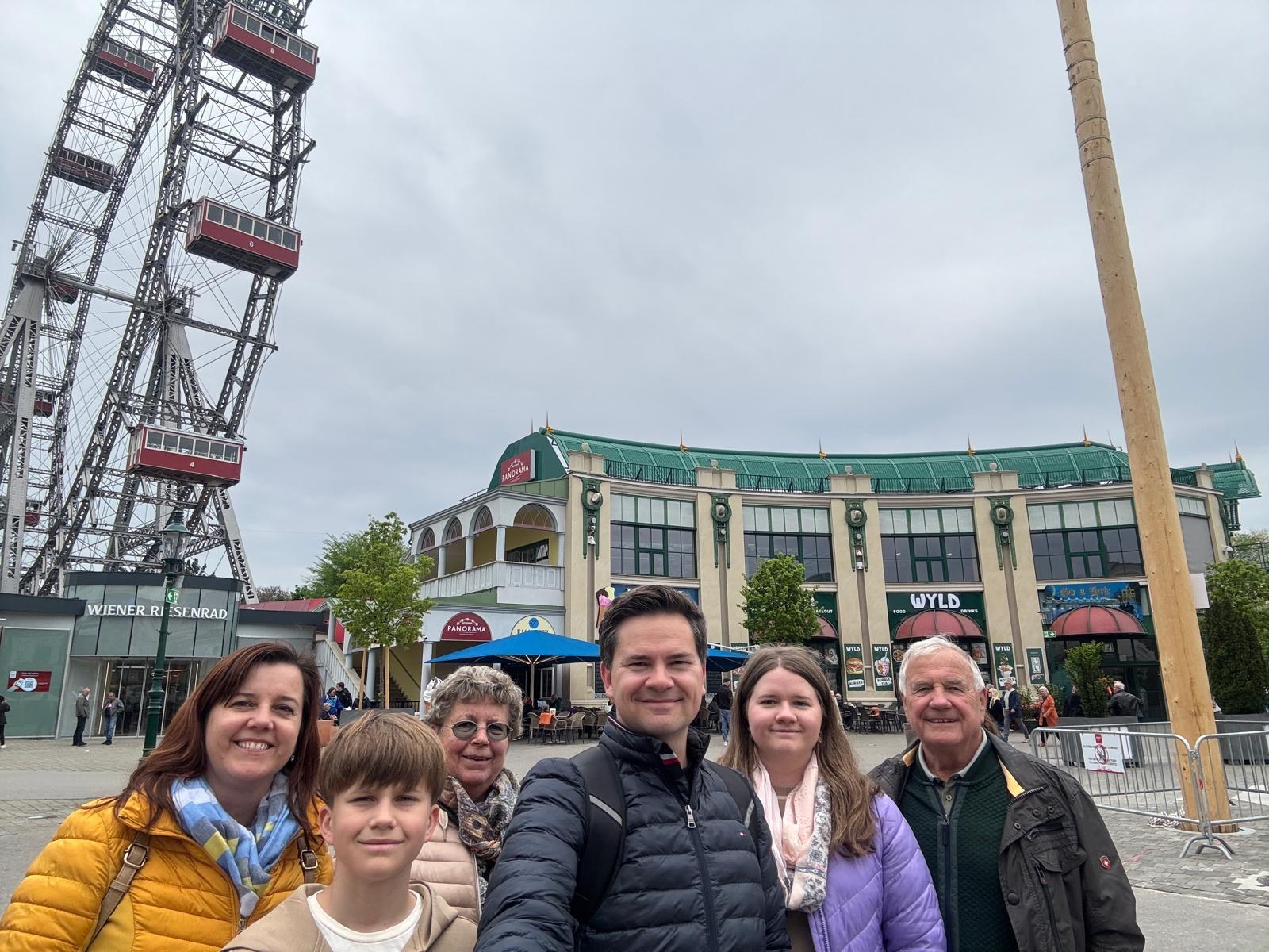 Eine Gruppe von sechs Personen macht ein Selfie im Freien vor einem großen Riesenrad. Im Vordergrund stehen ein Mann mit ausgestrecktem Arm, der das Foto aufnimmt, sowie zwei Kinder und drei ältere Erwachsene, alle lächeln in die Kamera. Im Hintergrund ist das bekannte Wiener Riesenrad sowie ein Gebäude mit grüner Dachkonstruktion und mehreren Geschäften zu sehen, unter einem grauen, bewölkten Himmel.