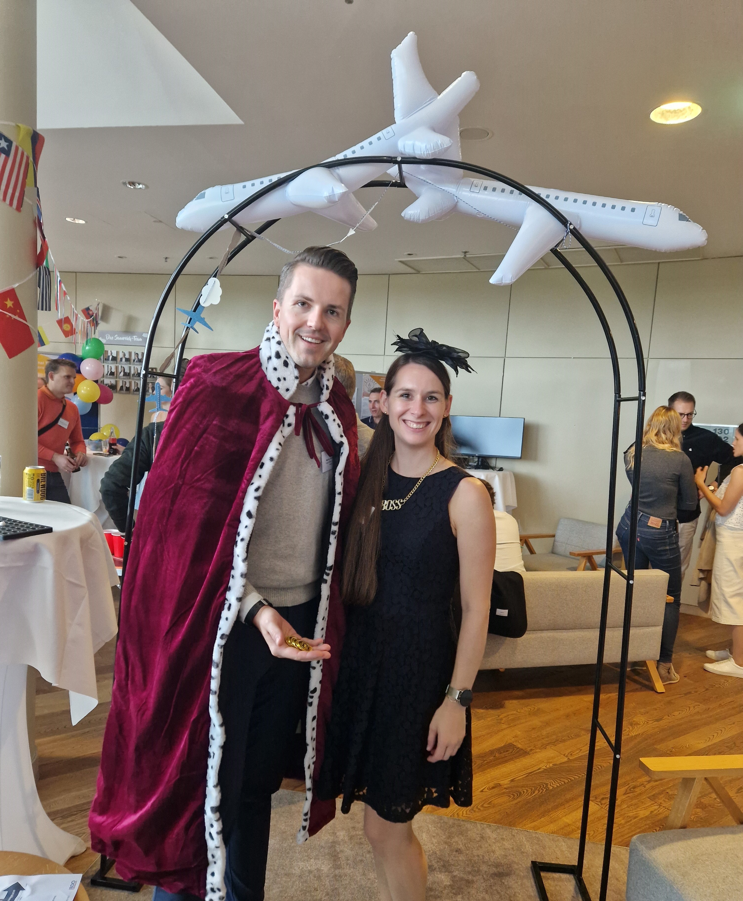 Zwei Personen in Kostüm (König und Lady) posieren unter einem Deko-Bogen mit aufblasbarem Flugzeug, im Hintergrund Büroparty mit internationalen Flaggen.