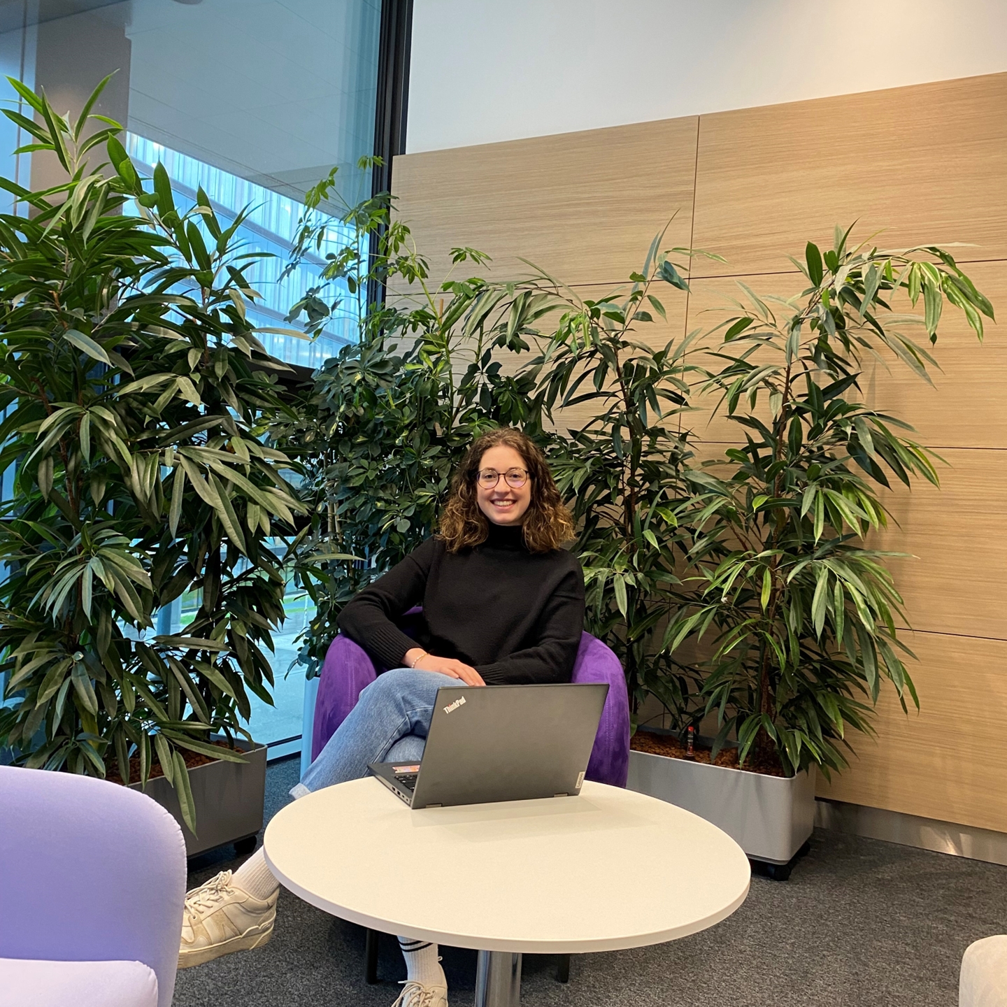 Eine junge Frau mit lockigem Haar und Brille sitzt entspannt in einem lila Sessel in einem modernen Büro, umgeben von großen grünen Pflanzen. Sie trägt einen schwarzen Pullover, Jeans und weiße Sneakers. Vor ihr auf einem kleinen runden Tisch steht ein aufgeklappter Laptop, und sie lächelt in die Kamera.