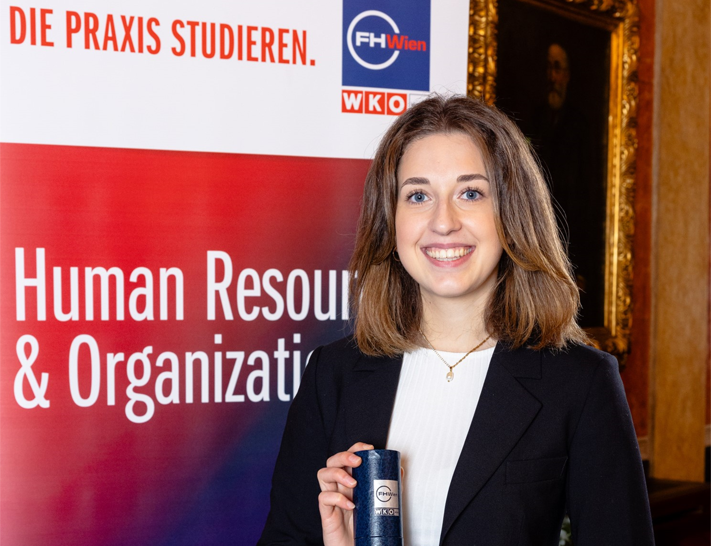 Eine junge Frau steht lächelnd vor einem Roll-up mit der Aufschrift „Human Resource & Organization“ der FH Wien. Sie hält eine gerollte Urkunde in der Hand und trägt einen dunklen Blazer.