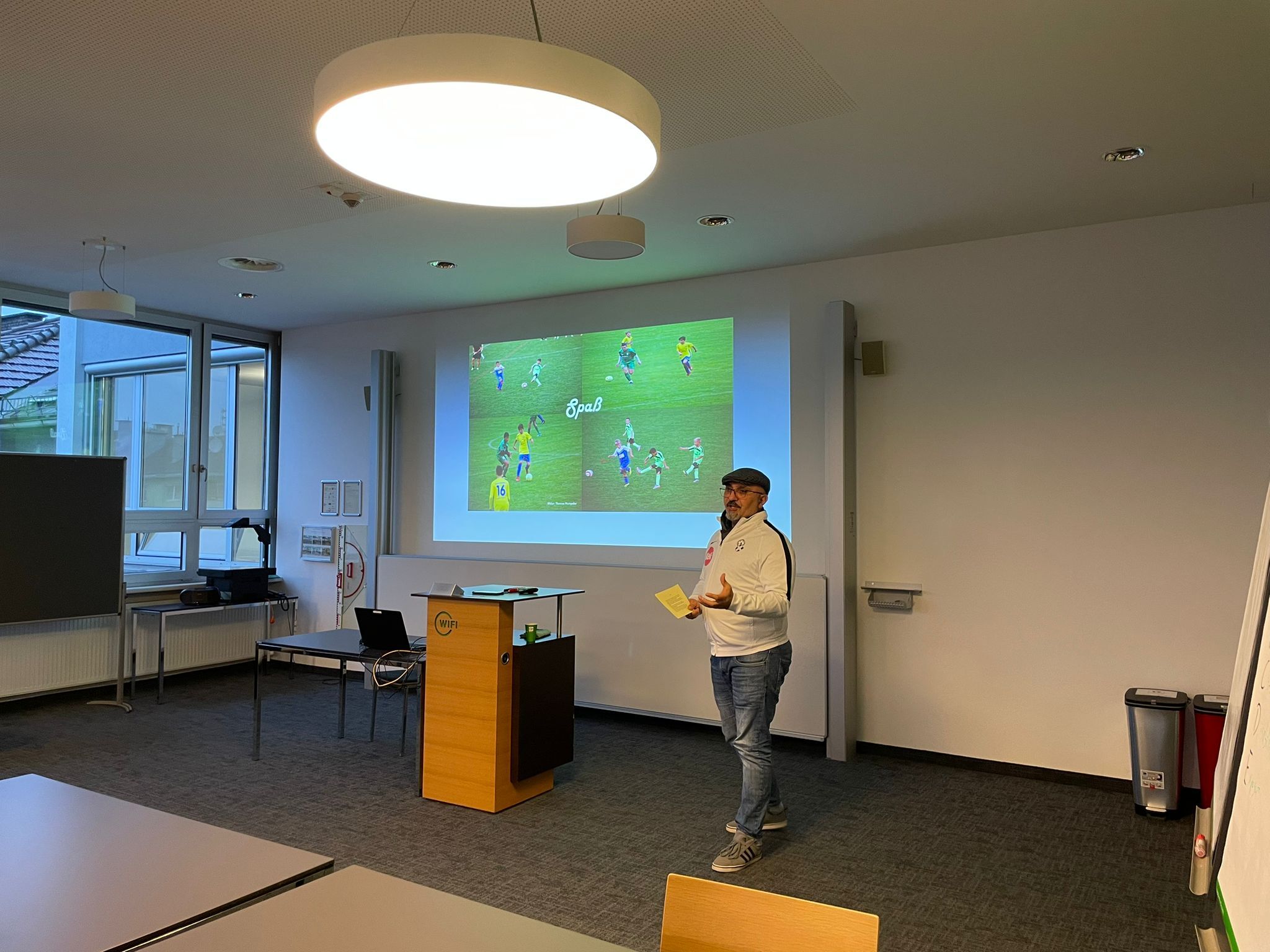 Mann bei einer Präsentation in einem Seminarraum, im Hintergrund eine Projektion mit Bildern zum Thema Sport