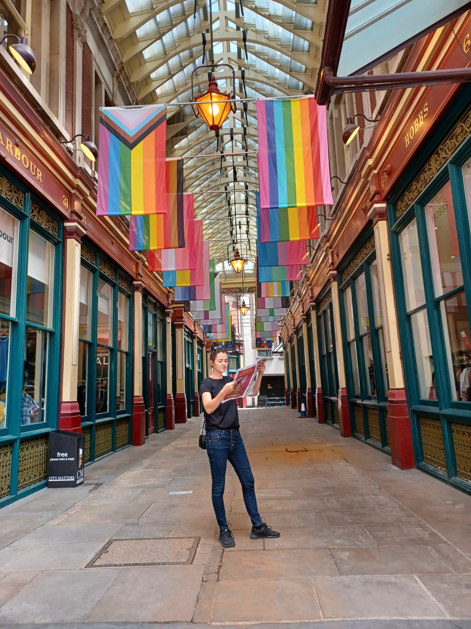 Eine Frau steht in einer überdachten Passage und liest eine Zeitschrift. Über ihr hängen zahlreiche Regenbogen- und Pride-Flaggen. Die Passage hat große Fenster und dekorative Fassaden, und Laternen hängen von der Decke. Die Frau trägt schwarze Kleidung und steht entspannt in der Mitte des Gangs.