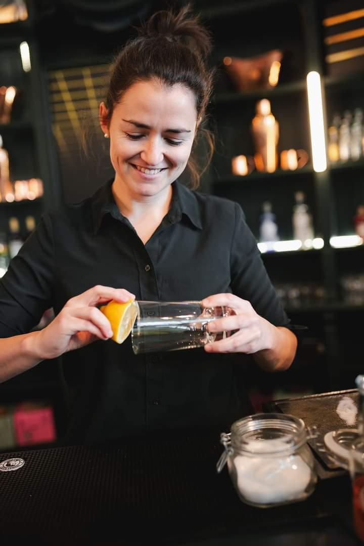 Eine Frau in schwarzer Kleidung steht hinter einer Bar und reibt den Rand eines Glases mit einer Zitronenscheibe ein. Sie lächelt dabei und wirkt konzentriert und zufrieden. Im Hintergrund sind Barregale mit Flaschen und Kupferdekorationen zu sehen.
