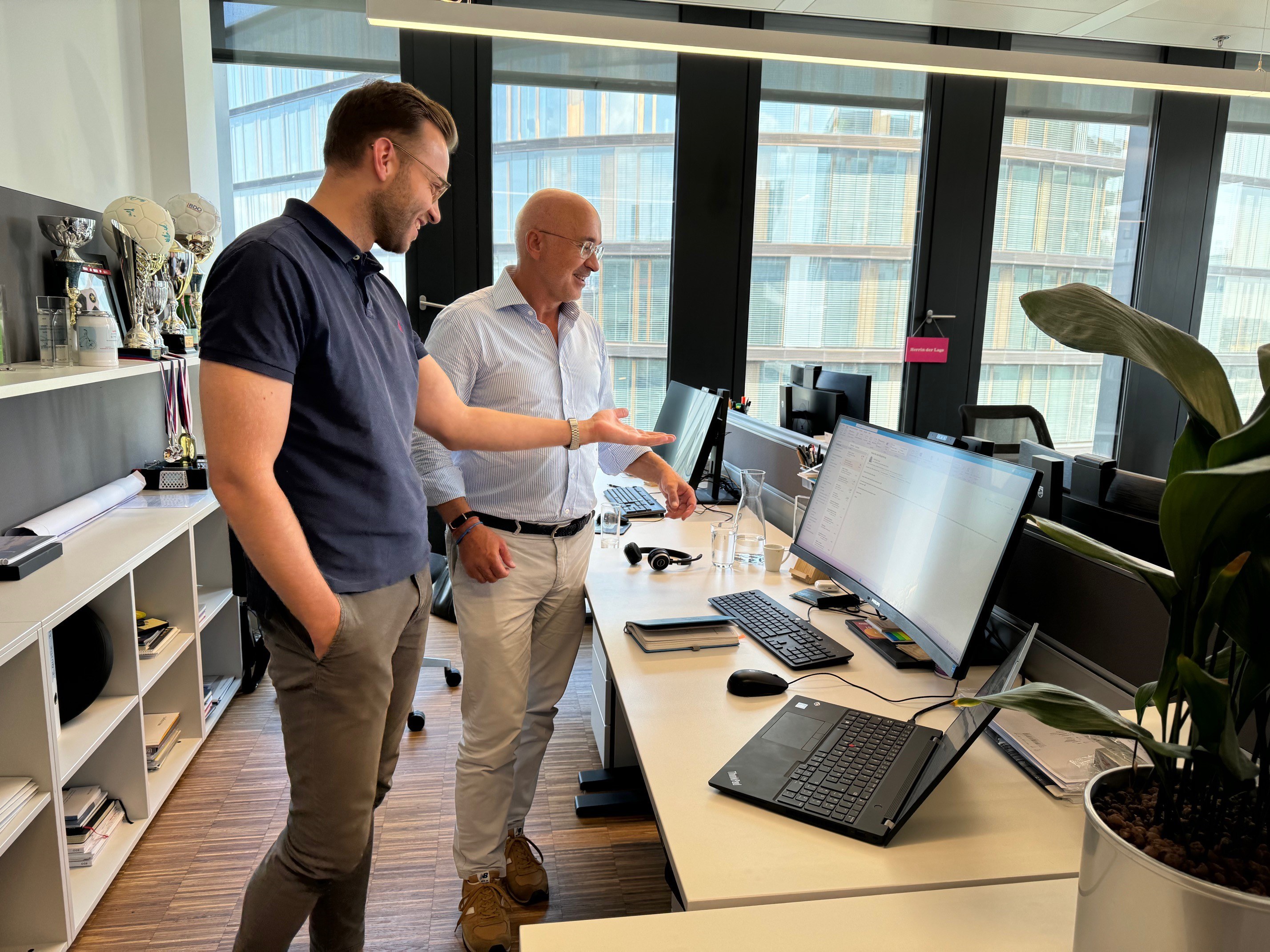 Zwei Männer stehen im modernen Büro vor mehreren Bildschirmen und unterhalten sich angeregt, Pokale und Medaillen im Hintergrund.