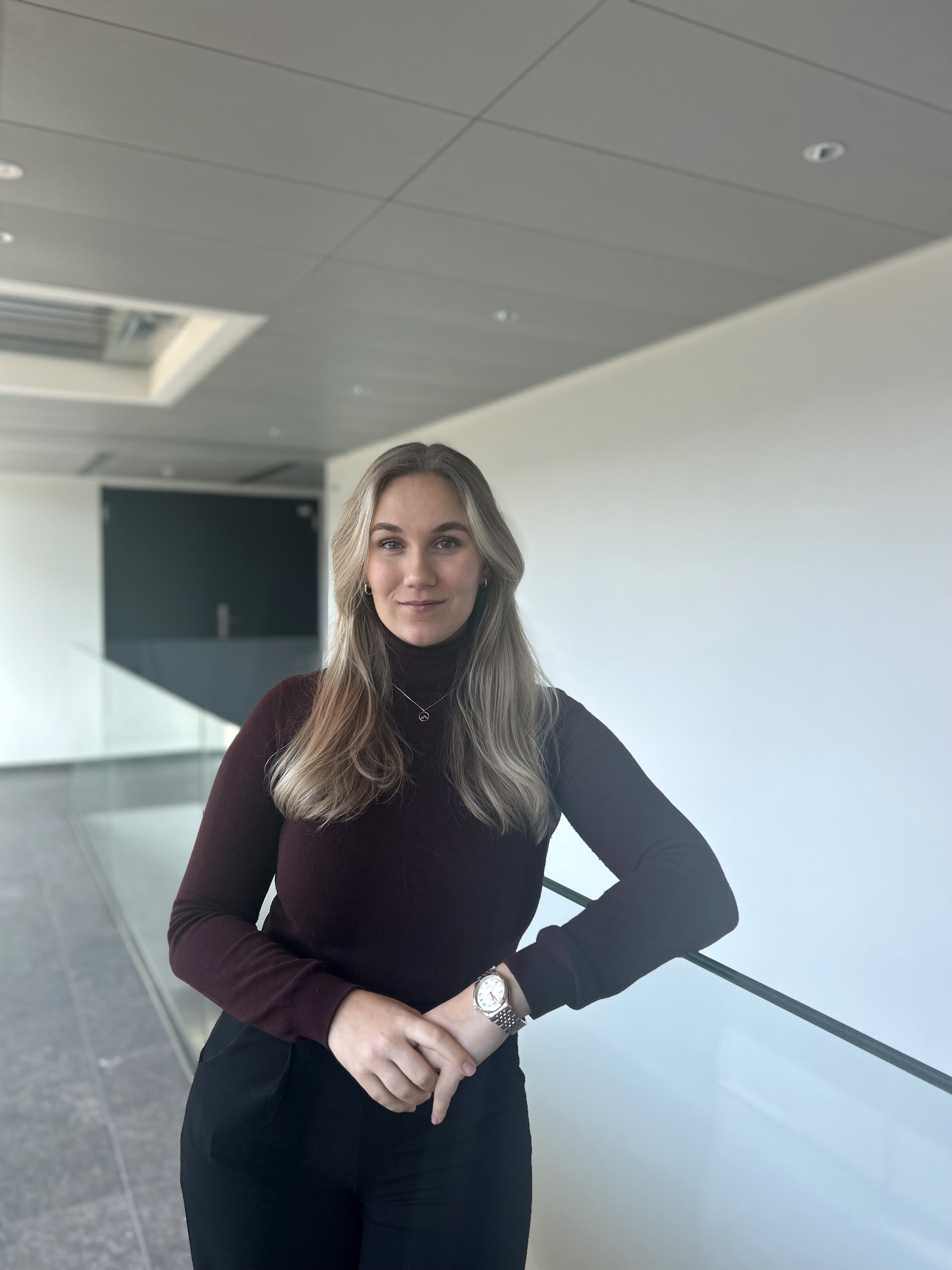Eine junge Frau mit langen, blonden Haaren steht in einem modernen, minimalistischen Bürogebäude. Sie trägt einen dunkelroten Rollkragenpullover und eine schwarze Hose. An ihrem Handgelenk trägt sie eine Armbanduhr, und sie blickt ruhig und selbstbewusst in die Kamera. Der Hintergrund ist hell und schlicht.