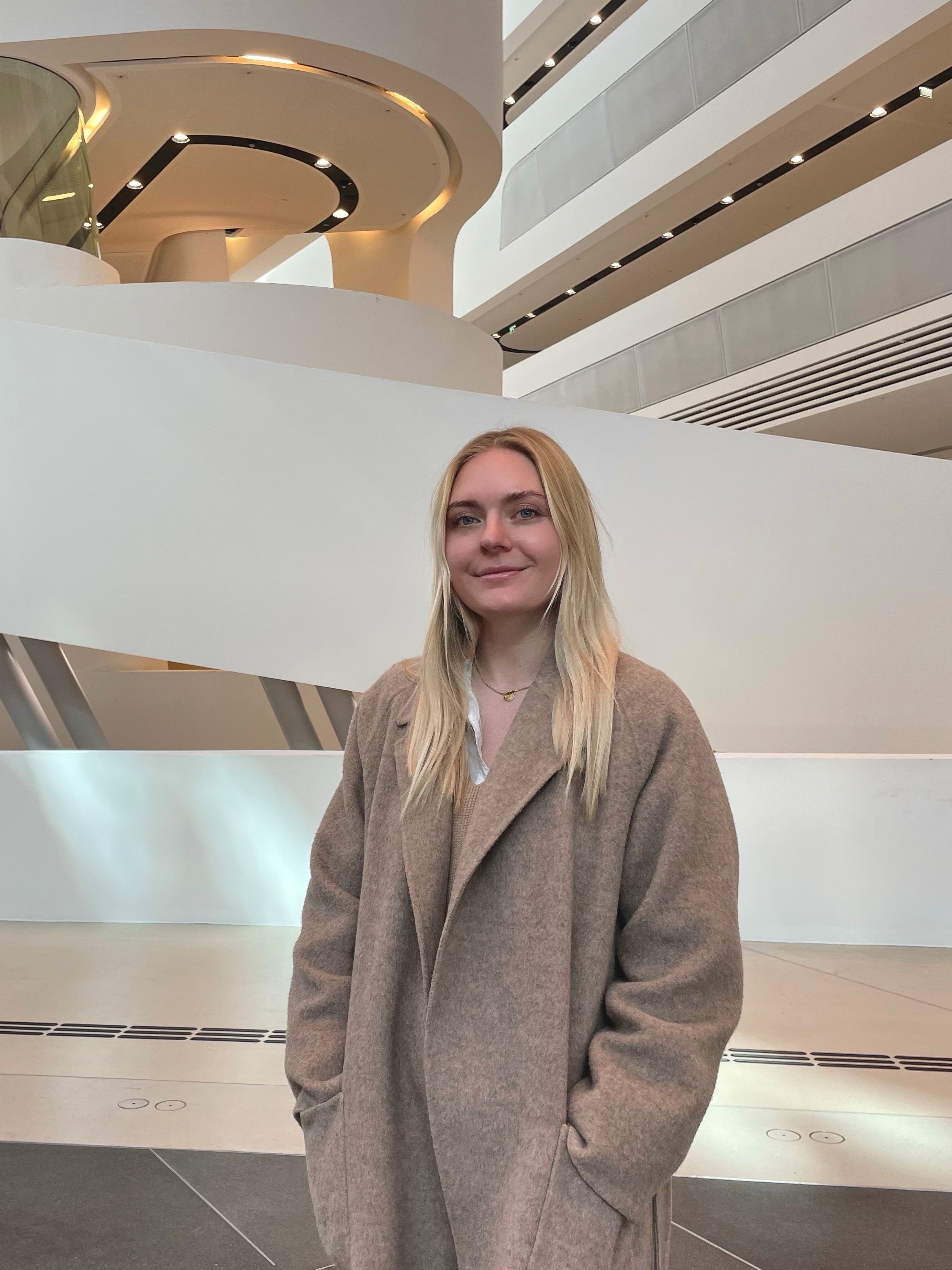 Eine junge Frau mit langen, blonden Haaren steht in einem modernen, futuristischen Gebäude (Wirtschaftsuniversität Wien) mit geschwungenen weißen Wänden und architektonischen Elementen. Sie trägt einen beigen Mantel und lächelt dezent in die Kamera. Der Raum ist groß und minimalistisch gestaltet.