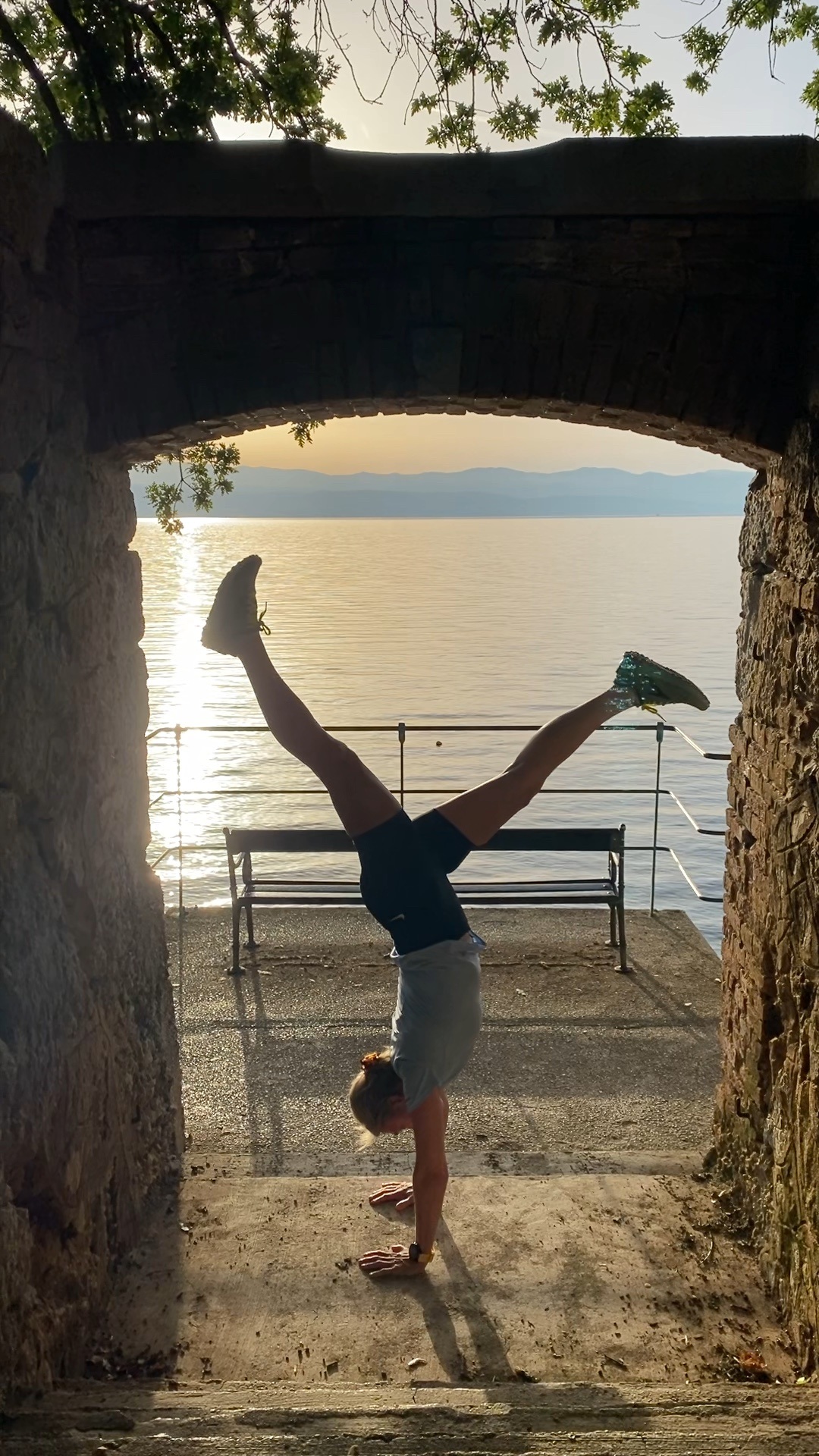 Eine Person macht einen Handstand unter einem steinernen Torbogen mit Blick auf einen Sonnenuntergang über einem ruhigen See.
