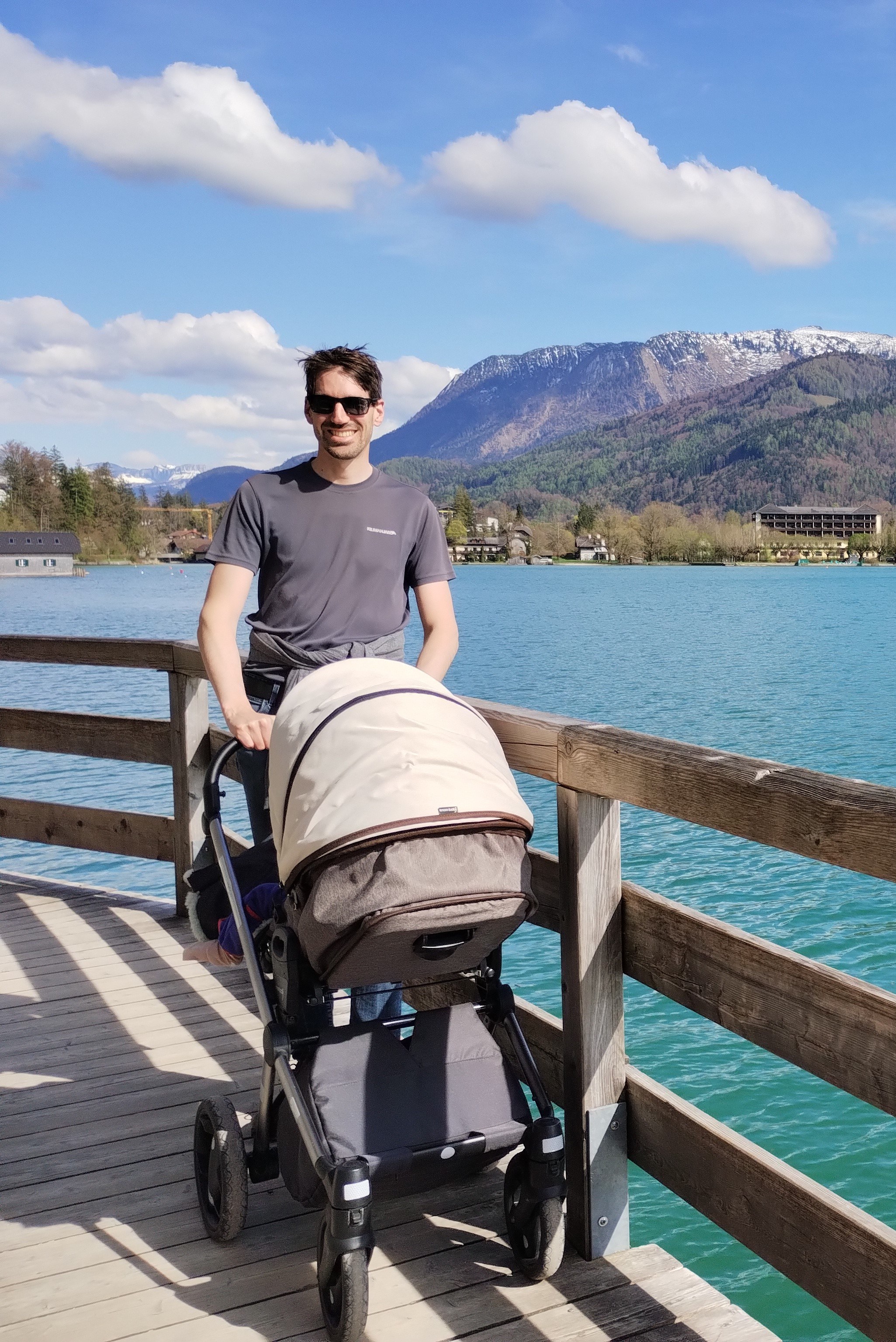 Ein Mann in Sonnenbrille steht auf einem Holzsteg an einem klaren See und schiebt einen Kinderwagen. Im Hintergrund sind bewaldete Hügel und schneebedeckte Berge unter einem blauen Himmel mit einigen Wolken zu sehen. Der Mann lächelt und genießt die Aussicht.