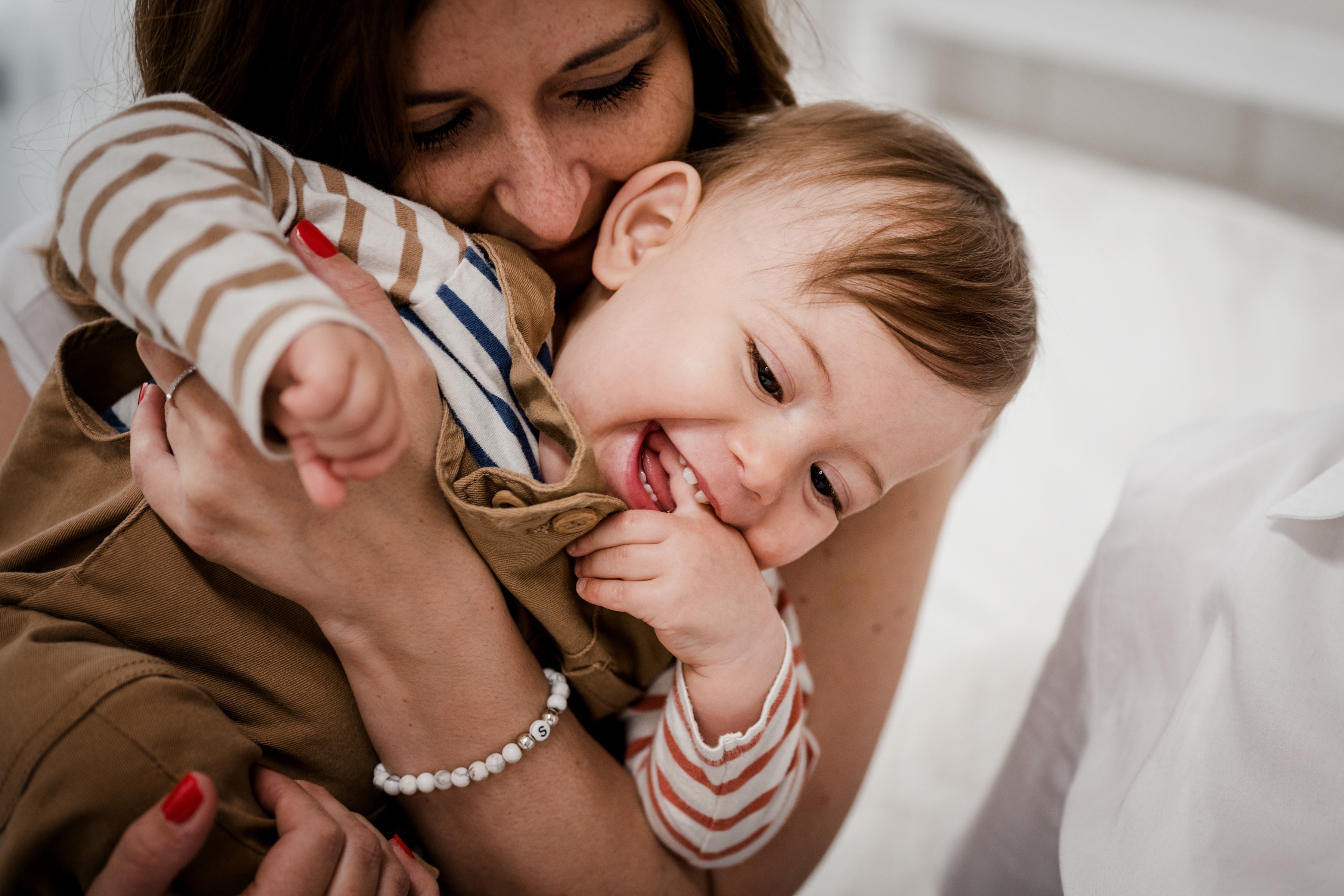 Eine Frau hält liebevoll ein lachendes Baby in ihren Armen. Das Baby trägt ein gestreiftes Langarmshirt und eine braune Latzhose. Die Frau drückt das Kind sanft an sich, während sie lächelt und die Nähe genießt. Beide wirken glücklich und entspannt.