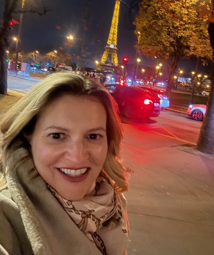 Lächelnde Frau macht ein Selfie bei Nacht in Paris, im Hintergrund ist der beleuchtete Eiffelturm zu sehen.