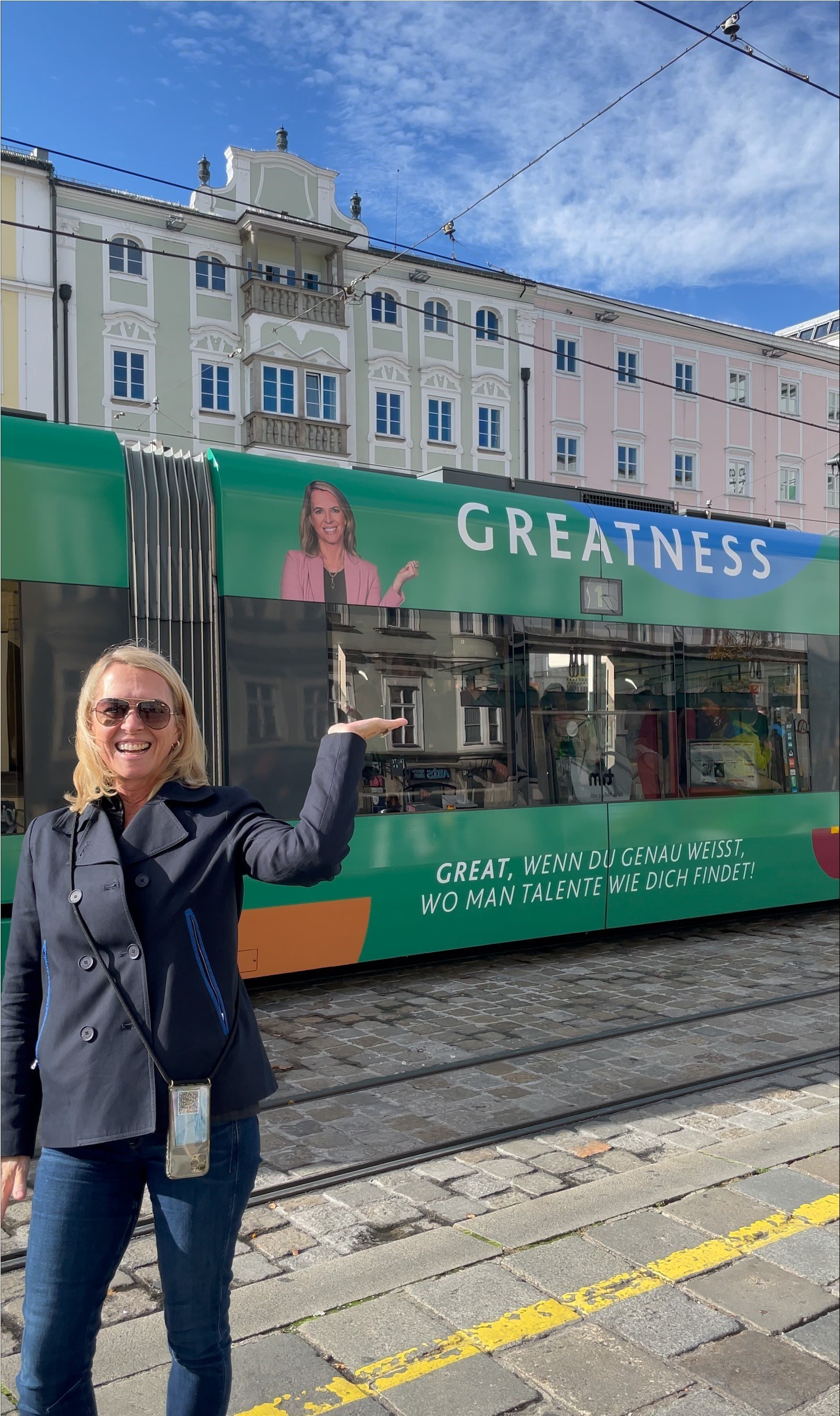Frau in dunkler Jacke steht lächelnd vor einer grünen Straßenbahn mit dem Wort "GREATNESS" und zeigt auf ein Werbebild mit ihrem eigenen Porträt.