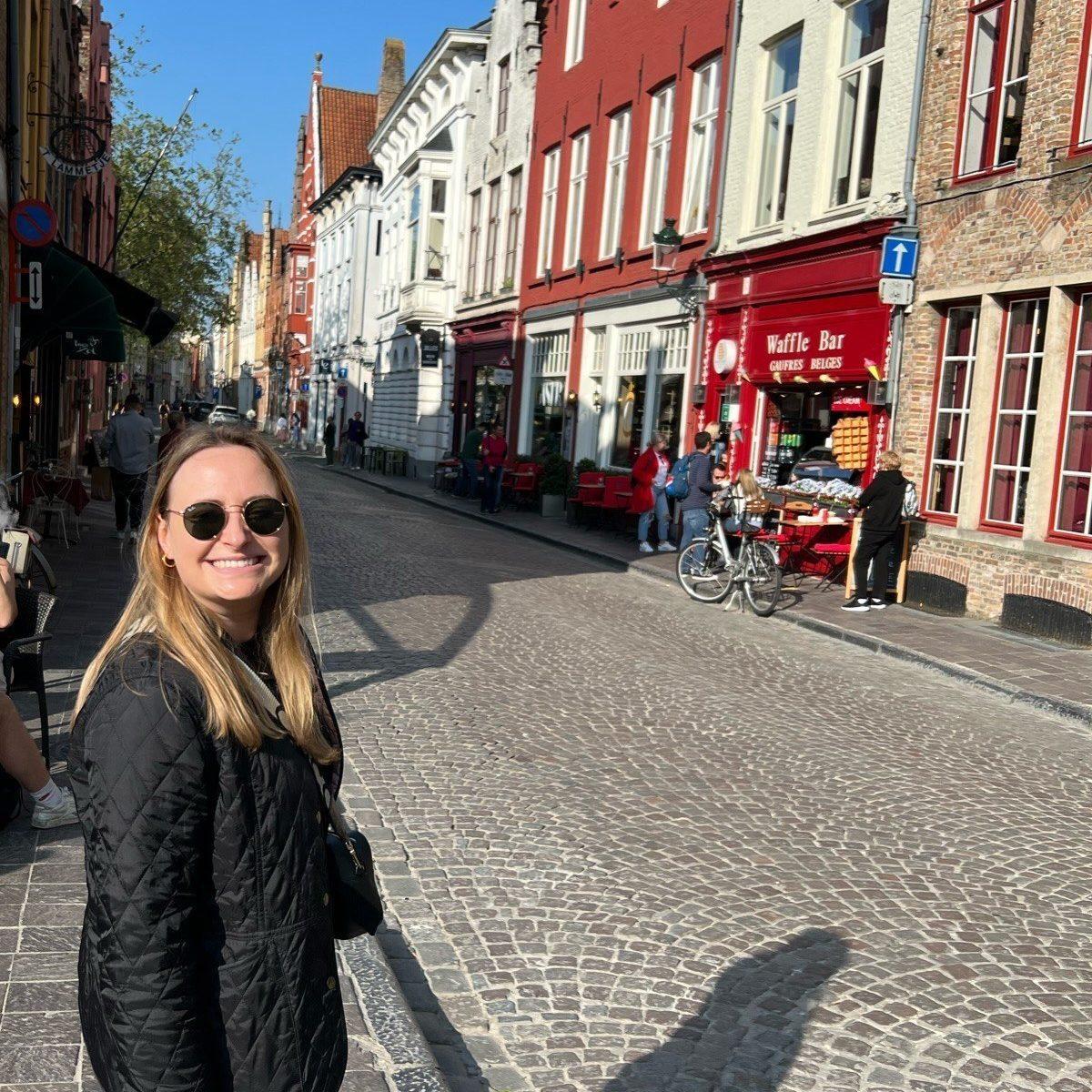 Eine lächelnde Frau steht auf einer sonnigen Kopfsteinpflasterstraße mit bunten Altstadthäusern im Hintergrund.