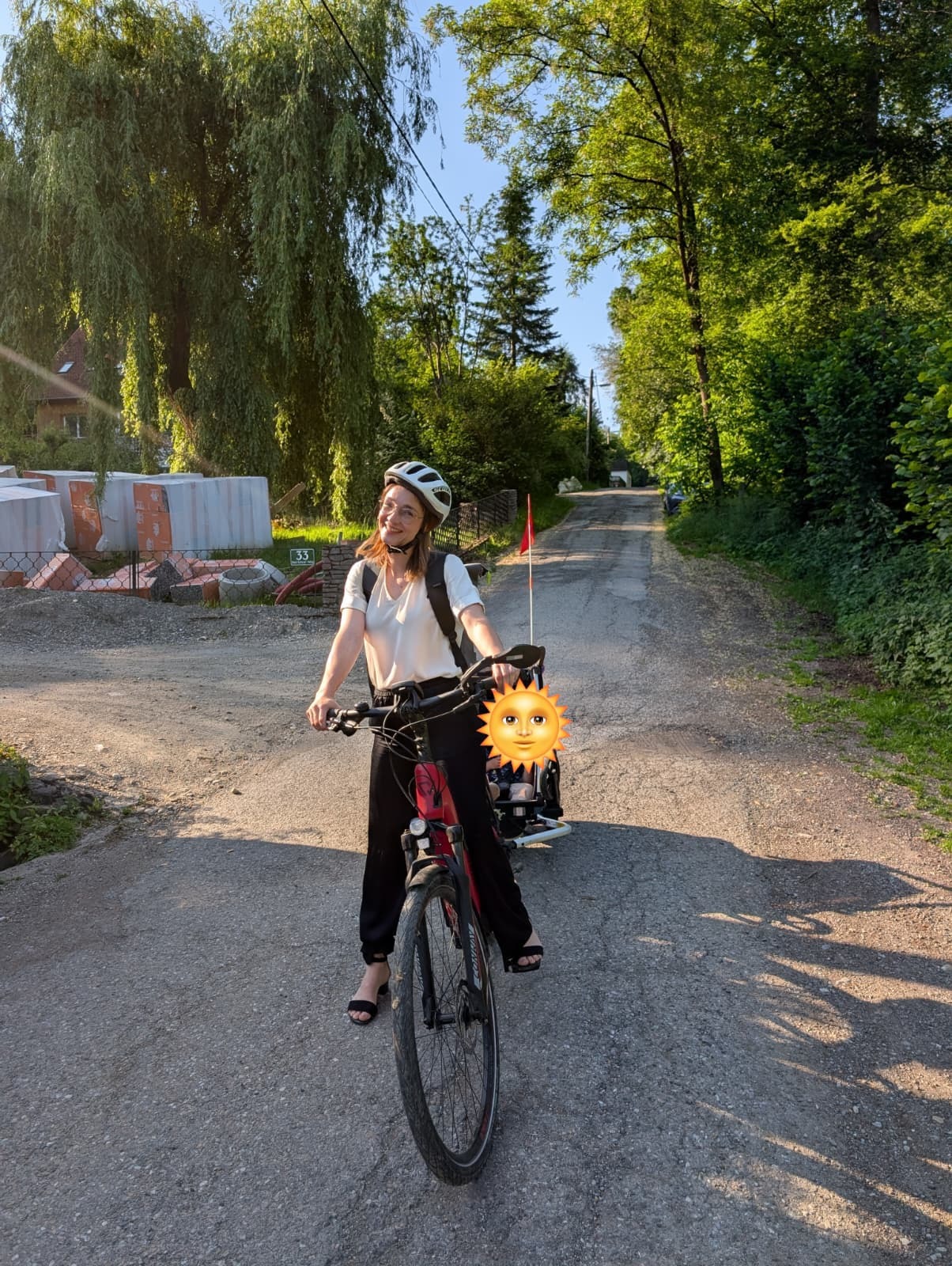 Eine lächelnde Frau mit Fahrradhelm steht mit ihrem Fahrrad auf einer ruhigen, von Bäumen gesäumten Straße. Am Fahrrad ist ein Kindersitz mit einem Kind befestigt, dessen Gesicht durch ein Sonnen-Emoji verdeckt ist. Die sommerliche Umgebung ist von üppigem Grün und warmem Sonnenlicht geprägt.