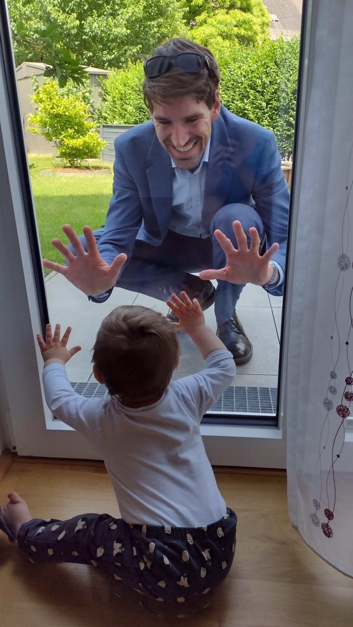 Ein Mann in einem blauen Anzug hockt draußen vor einer Glastür und lächelt, während er seine Hände gegen die Scheibe drückt. Ein kleines Kind sitzt drinnen und berührt mit den Händen die Glastür von der Innenseite, als würde es dem Mann entgegenkommen. Im Hintergrund ist ein grüner Garten zu sehen.