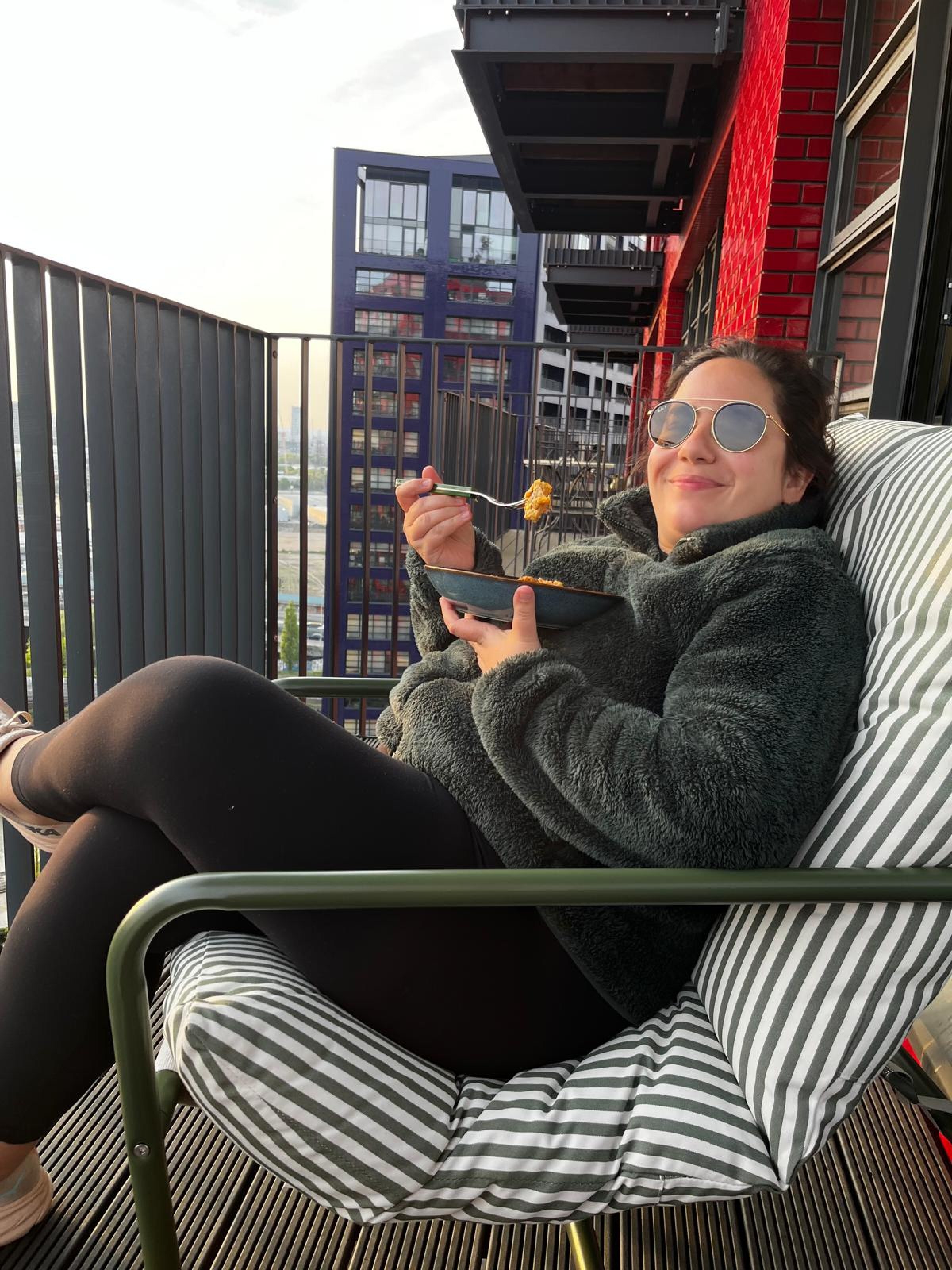 Frau mit Sonnenbrille sitzt entspannt auf einem Balkon und isst aus einer Schüssel.