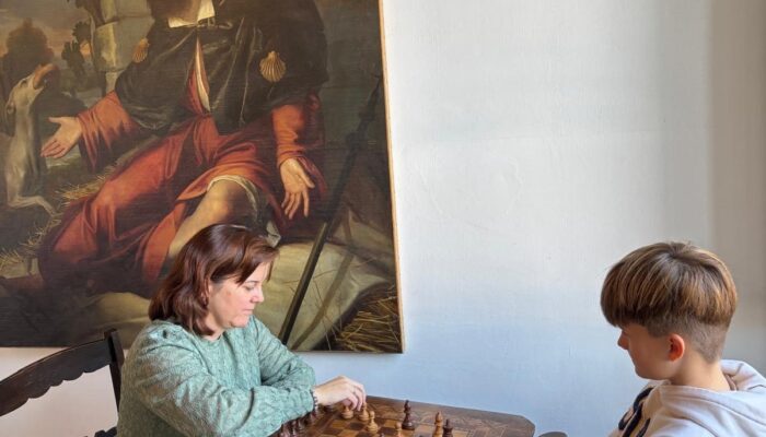 Ein Junge und eine Frau sitzen sich an einem kleinen Holztisch gegenüber und spielen konzentriert Schach. Die Frau trägt einen grünen Pullover und greift gerade nach einer Figur, während der Junge in einem hellen Hoodie aufmerksam zuschaut. Im Hintergrund hängt ein großes Gemälde mit einer religiösen Szene, das die ruhige, fast museale Atmosphäre des Raumes unterstreicht.