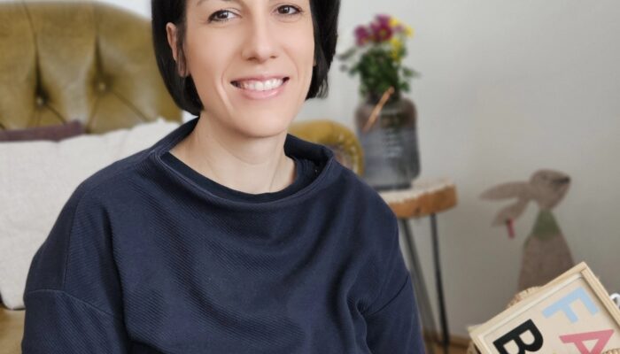 Eine Frau mit kurzen dunklen Haaren hält eine Tasse Kaffee in der Hand und lächelt.