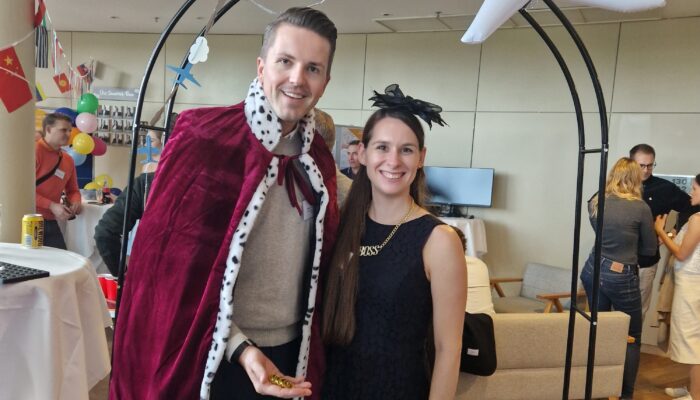 Zwei Personen in Kostüm (König und Lady) posieren unter einem Deko-Bogen mit aufblasbarem Flugzeug, im Hintergrund Büroparty mit internationalen Flaggen.