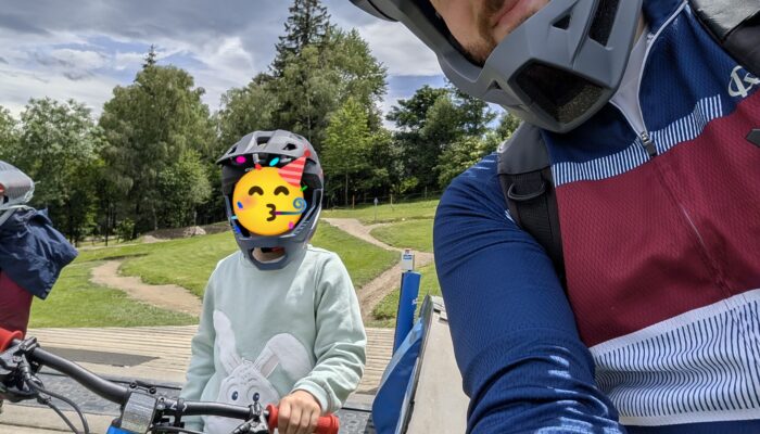 Selfie von einer Mountainbike-Tour: Eine Person im Fahrradhelm lächelt in die Kamera, während ein Kind mit Mountainbike und Helm daneben auf einer Holzplattform steht. Dahinter erstrecken sich eine grüne Wiese, Bäume und ein dramatisch bewölkter Himmel.