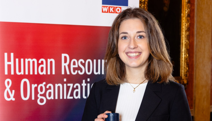 Eine junge Frau steht lächelnd vor einem Roll-up mit der Aufschrift „Human Resource & Organization“ der FH Wien. Sie hält eine gerollte Urkunde in der Hand und trägt einen dunklen Blazer.