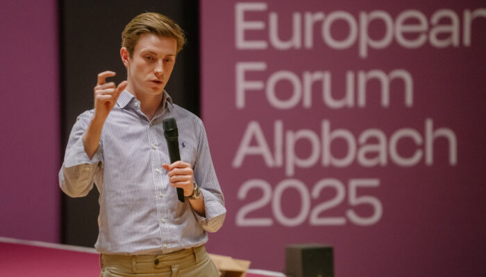 Ein junger Mann steht auf einer Bühne, hält ein Mikrofon in der Hand und gestikuliert beim Sprechen. Im Hintergrund ist eine große Leinwand mit der Aufschrift „European Forum Alpbach 2025“ zu sehen.