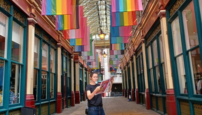 Eine Frau steht in einer überdachten Passage und liest eine Zeitschrift. Über ihr hängen zahlreiche Regenbogen- und Pride-Flaggen. Die Passage hat große Fenster und dekorative Fassaden, und Laternen hängen von der Decke. Die Frau trägt schwarze Kleidung und steht entspannt in der Mitte des Gangs.
