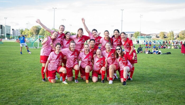 Eine jubelnde Frauenfußballmannschaft steht auf einem Fußballfeld. Sie tragen rote Fußballdressen.