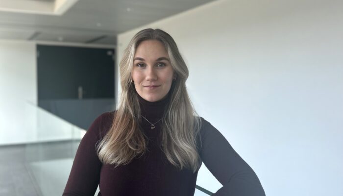 Eine junge Frau mit langen, blonden Haaren steht in einem modernen, minimalistischen Bürogebäude. Sie trägt einen dunkelroten Rollkragenpullover und eine schwarze Hose. An ihrem Handgelenk trägt sie eine Armbanduhr, und sie blickt ruhig und selbstbewusst in die Kamera. Der Hintergrund ist hell und schlicht.