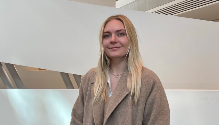 Eine junge Frau mit langen, blonden Haaren steht in einem modernen, futuristischen Gebäude (Wirtschaftsuniversität Wien) mit geschwungenen weißen Wänden und architektonischen Elementen. Sie trägt einen beigen Mantel und lächelt dezent in die Kamera. Der Raum ist groß und minimalistisch gestaltet.
