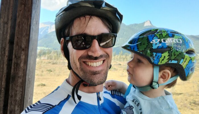 Mann mit Fahrradhelm und Sonnenbrille hält ein kleines Kind im Arm, beide tragen Helme und lächeln vor einer Berglandschaft bei sonnigem Wetter.