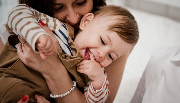 Eine Frau hält liebevoll ein lachendes Baby in ihren Armen. Das Baby trägt ein gestreiftes Langarmshirt und eine braune Latzhose. Die Frau drückt das Kind sanft an sich, während sie lächelt und die Nähe genießt. Beide wirken glücklich und entspannt.