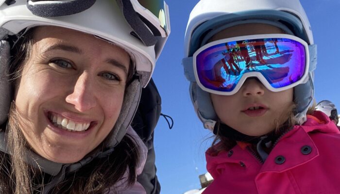 Frau und Kind mit Skibrillen und Helmen machen ein fröhliches Selfie auf der Skipiste vor blauem Himmel.