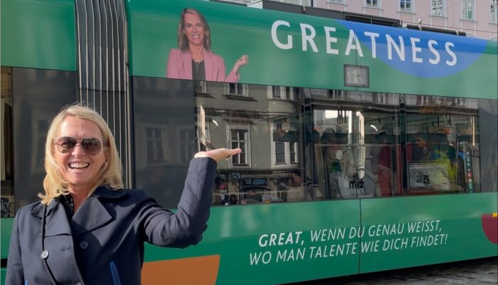 Frau in dunkler Jacke steht lächelnd vor einer grünen Straßenbahn mit dem Wort "GREATNESS" und zeigt auf ein Werbebild mit ihrem eigenen Porträt.