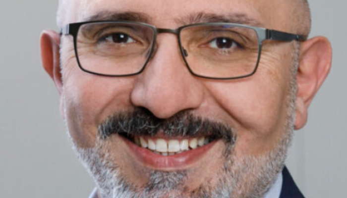 Porträt eines lächelnden Mannes mit Brille und grauem Bart im dunkelblauen Sakko vor hellem Hintergrund