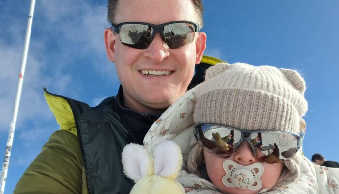Ein lächelnder Mann in Sonnenbrille hält ein Baby im Arm. Beide sind warm gekleidet in Winterjacken. Das Baby trägt eine Mütze, ebenfalls eine Sonnenbrille und einen Schnuller und hält ein kleines gelbes Plüschtier. Im Hintergrund ist ein blauer Himmel mit wenigen Wolken zu sehen.