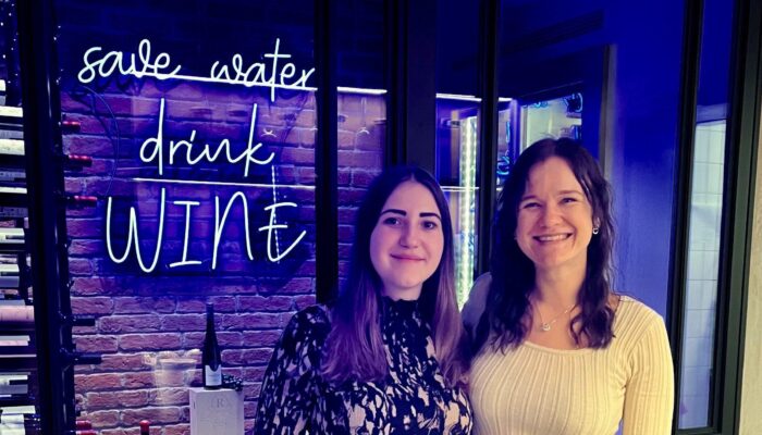 Zwei Kolleginnen posieren lächelnd in einer blau beleuchteten Weinbar mit Neon-Schriftzug im Hintergrund.